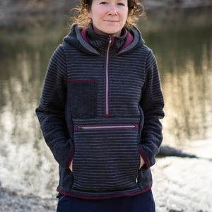 Könnte beinhalten: Ein grauer Kapuzenpullover mit horizontalen Streifen, einer Fronttasche und einem Viertel-Reißverschluss. Der Pullover hat bordeauxfarbene Verzierungen und wird mit einem marineblauen Rock kombiniert. Die Person steht im Freien in der Nähe eines Gewässers.