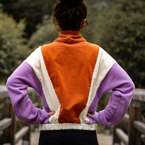 Puede incluir: Una sudadera de color bloque morado, blanco y naranja con cintura de cordón.