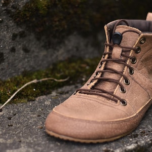 May include: A brown leather boot with a high-top design. The boot features a lace-up closure with brown laces, a padded collar, and a textured sole. The boot is set against a natural background with moss and stone.
