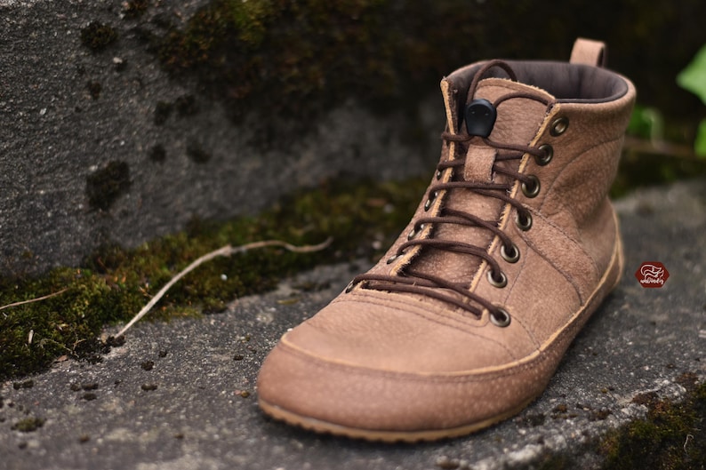 May include: A brown leather boot with a high-top design. The boot features a lace-up closure with brown laces, a padded collar, and a textured sole. The boot is set against a natural background with moss and stone.