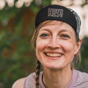 May include: A woman with long brown braids smiles while wearing a black and white patterned cycling cap with the word "HEY!" in white lettering.