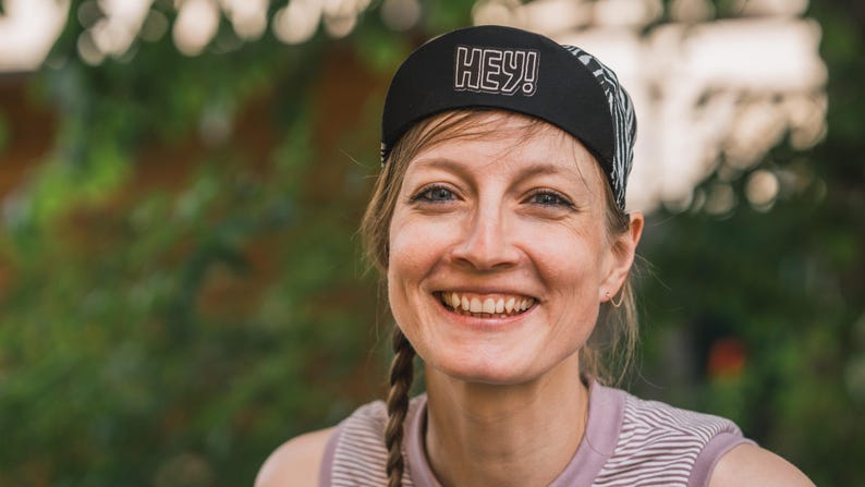 May include: A woman with long brown braids smiles while wearing a black and white patterned cycling cap with the word "HEY!" in white lettering.
