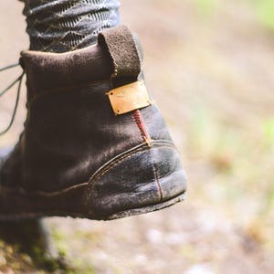 May include: Close-up of a worn, dark brown leather boot with a pull tab and a small gold-colored tag. The boot has a rugged sole and a red stitch detail on the heel. The laces are visible.