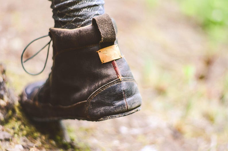 May include: Close-up of a worn, dark brown leather boot with a pull tab and a small gold-colored tag. The boot has a rugged sole and a red stitch detail on the heel. The laces are visible.