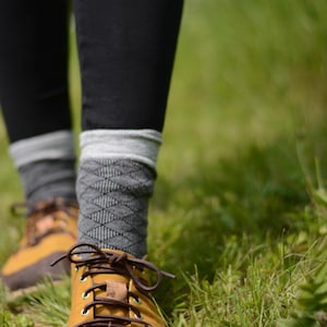 May include: Mustard yellow hiking boots with brown laces and a brown leather tongue. The boots are paired with gray patterned socks and black leggings. The image is taken outdoors in a grassy area.