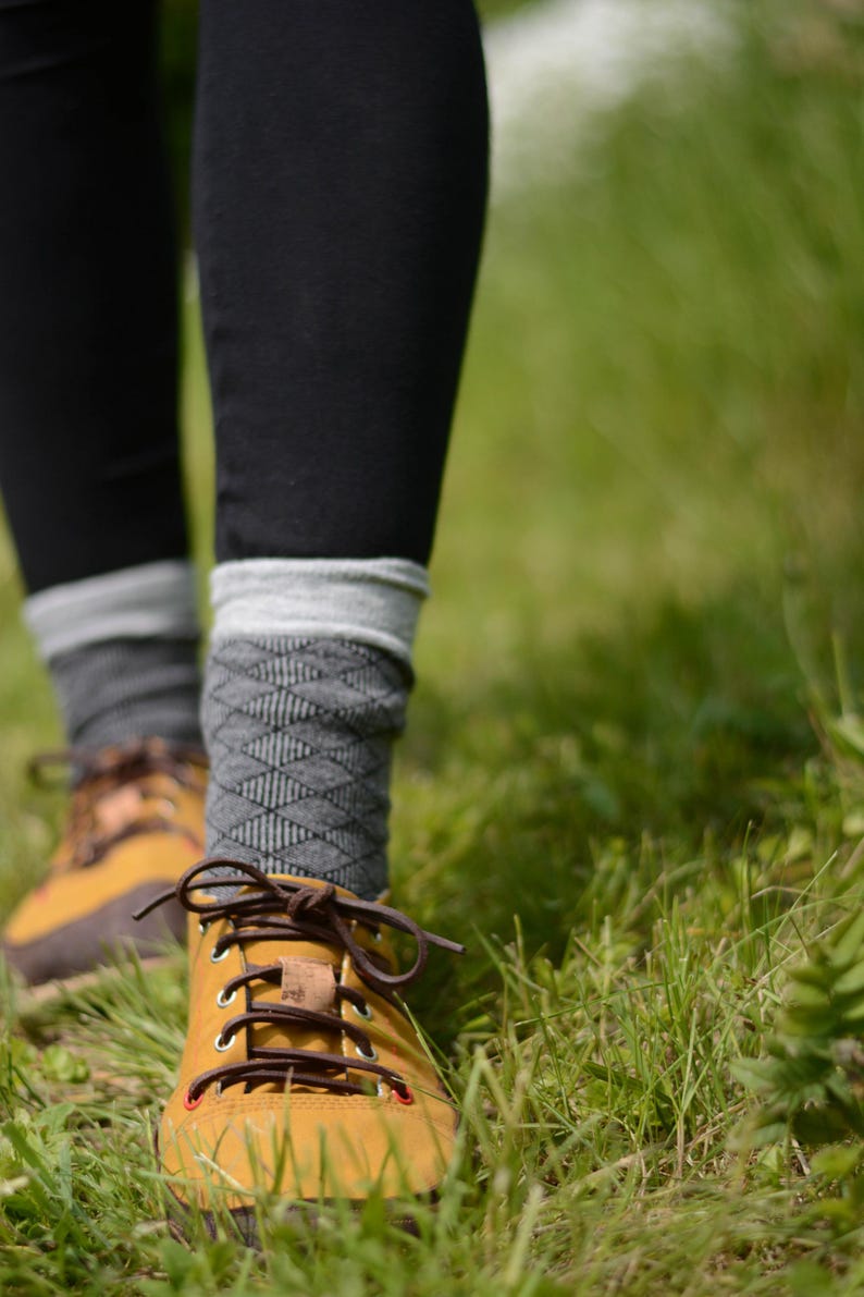 May include: Mustard yellow hiking boots with brown laces and a brown leather tongue. The boots are paired with gray patterned socks and black leggings. The image is taken outdoors in a grassy area.