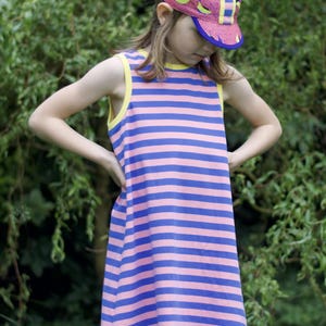 May include: A young person wearing a pink and blue striped sleeveless dress with a yellow trim. They are also wearing a pink and yellow patterned baseball cap.