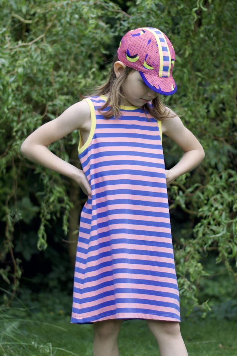 May include: A young person wearing a pink and blue striped sleeveless dress with a yellow trim. They are also wearing a pink and yellow patterned baseball cap.