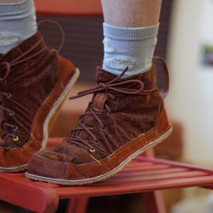 May include: Brown suede ankle boots with a lace-up design. The boots have a textured pattern and a contrasting orange sole. The laces and pull tabs are a matching brown. The boots are worn with light blue socks.
