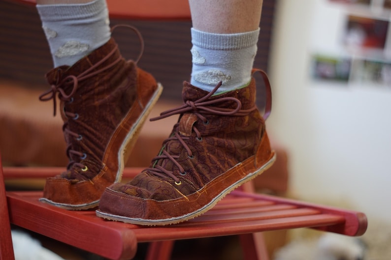 May include: Brown suede ankle boots with a lace-up design. The boots have a textured pattern and a contrasting orange sole. The laces and pull tabs are a matching brown. The boots are worn with light blue socks.