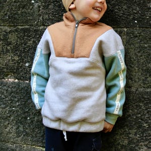 Puede incluir: Una sudadera gris claro con bloques de color marrón y azul. Las mangas tienen una franja azul claro y blanca con la palabra "happy" repetida.