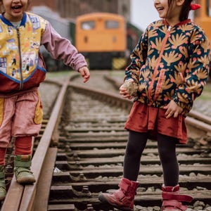 May include: Two children wearing colorful patterned jackets stand on a set of train tracks. The girl on the left is wearing a pink and yellow jacket with a whimsical design. The girl on the right is wearing a dark green jacket with a floral pattern. Both girls are smiling.
