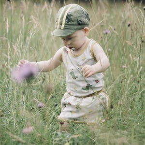 May include: A young child wearing a green and yellow patterned cap and a white tank top with a green and yellow pattern walks through a field of tall grass.