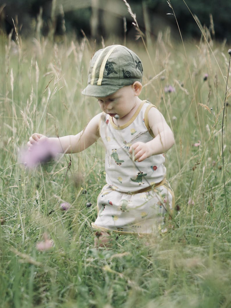 May include: A young child wearing a green and yellow patterned cap and a white tank top with a green and yellow pattern walks through a field of tall grass.