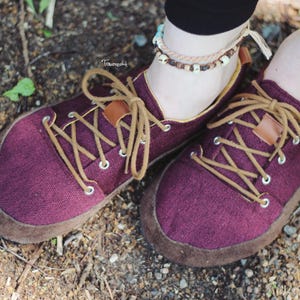 May include: Close-up of a pair of burgundy canvas shoes with brown suede soles and tan laces. The shoes have a minimalist design with a low profile and are suitable for casual wear. The laces are crisscrossed and secured through metal eyelets.