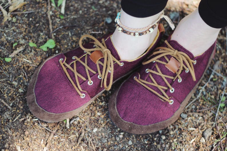 May include: Close-up of a pair of burgundy canvas shoes with brown suede soles and tan laces. The shoes have a minimalist design with a low profile and are suitable for casual wear. The laces are crisscrossed and secured through metal eyelets.