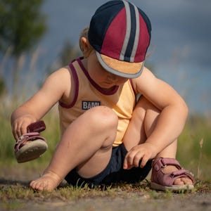 May include: A young child wearing a yellow tank top with the word "B&Auml;M!" on it, a blue and red striped cap, and pink sandals. The child is sitting on the ground and looking down at their feet.