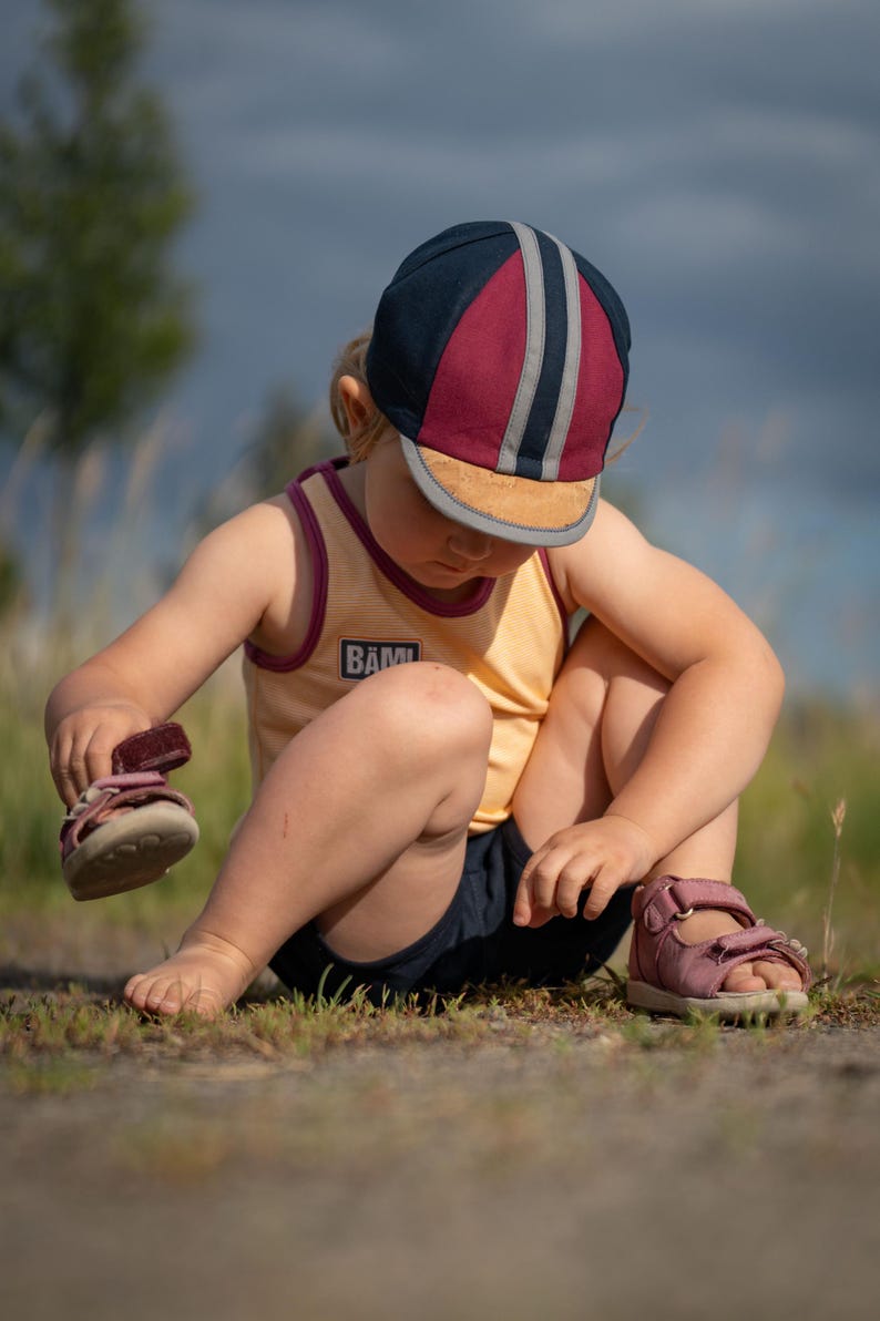 May include: A young child wearing a yellow tank top with the word "B&Auml;M!" on it, a blue and red striped cap, and pink sandals. The child is sitting on the ground and looking down at their feet.