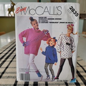 May include: A vintage McCall's sewing pattern featuring illustrations of three girls wearing oversized sweaters and leggings. The pattern includes the text "Easy McCall's" and "Focus on Overlock (Serger) Sewing". The pattern is for a girl's size medium.
