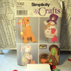 May include: A Simplicity Crafts sewing pattern envelope numbered 7002 featuring illustrations of four stuffed figures: an orange bear, a snowman with a purple hat, a rabbit in a colorful dress, and a green-faced character with a brown hat.