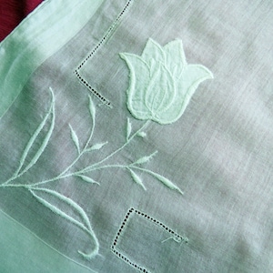 May include: A close-up of a white embroidered handkerchief with a floral design. The handkerchief features a large embroidered tulip and delicate stems and leaves. The edges are bordered with a light green trim and decorative stitching.