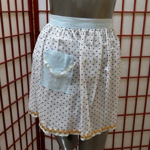 Puede incluir: Un delantal blanco con lunares negros, un bolsillo y una cintura azul claro. El bolsillo está adornado con encaje blanco. El borde inferior del delantal está adornado con encaje dorado.