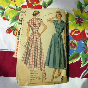 May include: A vintage sewing pattern envelope labeled 2842, size 14, bust 32, and 25 cents. The front features illustrations of two dresses: one with a red and white plaid pattern and another in solid blue, both with short sleeves and waist ties.