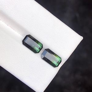 May include: Two emerald-cut, dark green gemstones with a polished finish. The gems are displayed on a white surface, showcasing their rectangular shape and facets. The stones have a deep, rich colour.