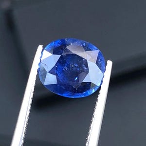 May include: A faceted, oval-cut sapphire gemstone. The sapphire is a deep, vibrant blue color and is held by silver tweezers against a dark background. The gemstone's facets reflect light, creating a sparkling effect.