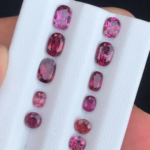 May include: A collection of oval and round gemstones in varying shades of pink and red. The gemstones are displayed on a white surface, showcasing their polished facets and vibrant colors. The gems vary in size.
