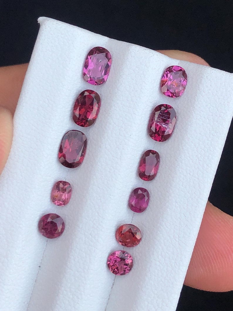 May include: A collection of oval and round gemstones in varying shades of pink and red. The gemstones are displayed on a white surface, showcasing their polished facets and vibrant colors. The gems vary in size.