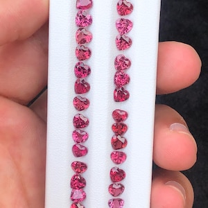 May include: Rows of heart-shaped gemstones in varying shades of red and pink are displayed on a white surface. The gemstones are arranged in vertical columns, showcasing their polished facets and vibrant colors. The image highlights the gemstones' romantic shape.