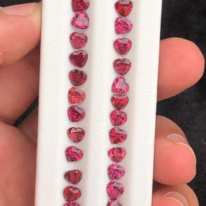 May include: Rows of heart-shaped gemstones in varying shades of red and pink are displayed on a white background. The gemstones are arranged in vertical columns, showcasing their facets and color variations. The image highlights the gemstones' cut and color.