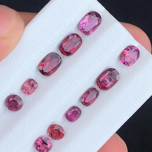 May include: A collection of oval and round-cut gemstones in varying shades of pink and red. The gemstones are displayed on a white surface, showcasing their polished facets and internal clarity. The gems are arranged in rows, highlighting their different sizes and shapes.