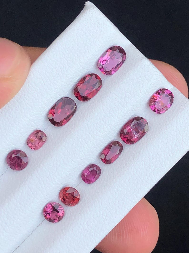 May include: A collection of oval and round-cut gemstones in varying shades of pink and red. The gemstones are displayed on a white surface, showcasing their polished facets and internal clarity. The gems are arranged in rows, highlighting their different sizes and shapes.