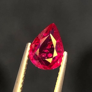 Puede incluir: Un rubí facetado en forma de pera, sujeto por unas pinzas doradas. El rubí muestra un tono rojo intenso con reflejos dorados internos. El corte de la gema revela múltiples facetas, realzando su brillo sobre un fondo oscuro.