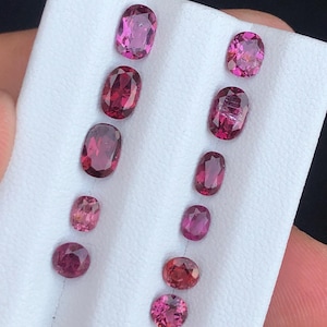 May include: A collection of oval and round gemstones in varying shades of pink and red, displayed on a white surface. The gemstones are of different sizes and shapes, showcasing their natural beauty and color variations.