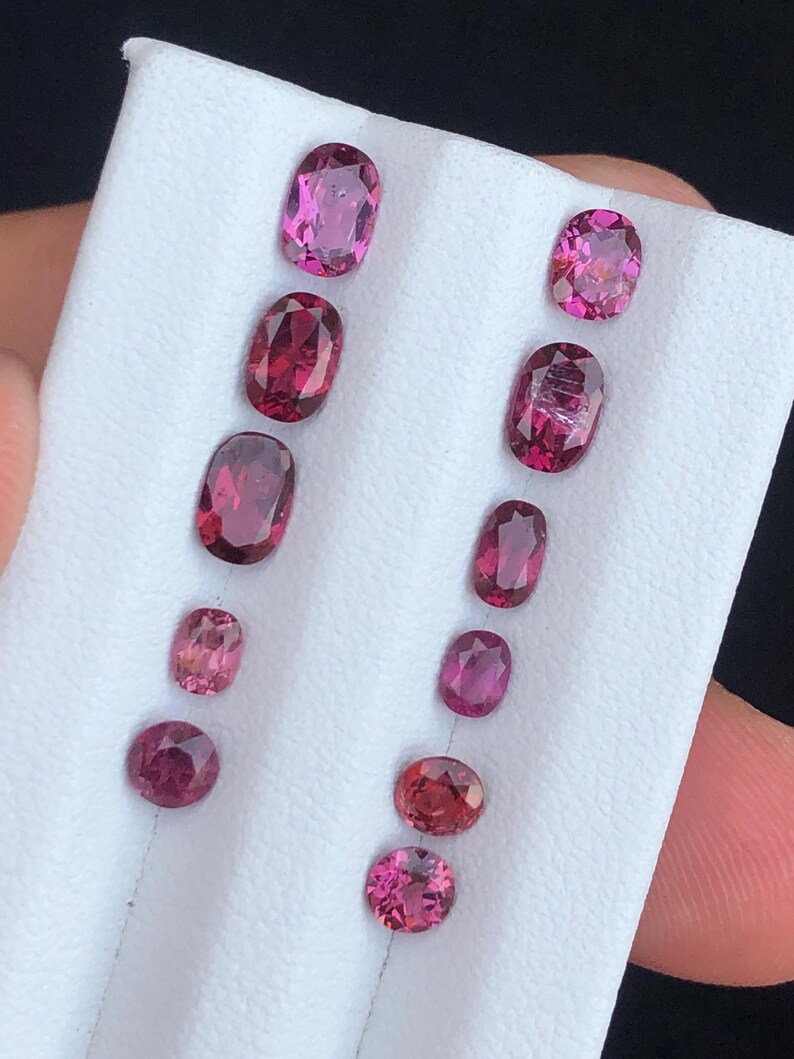 May include: A collection of oval and round gemstones in varying shades of pink and red, displayed on a white surface. The gemstones are of different sizes and shapes, showcasing their natural beauty and color variations.