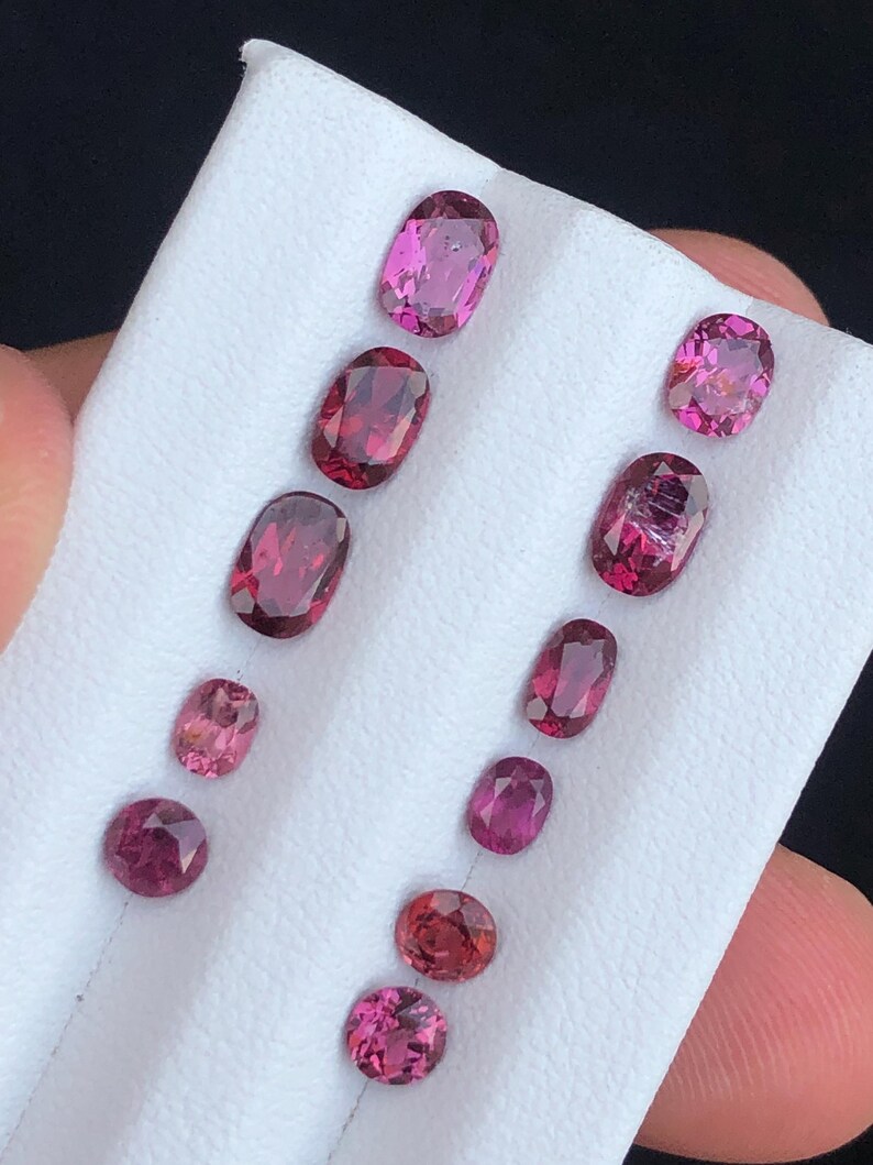 May include: A collection of oval and round gemstones in varying shades of pink and red, displayed on a white surface. The gemstones are faceted, showcasing their internal structure and brilliance. The arrangement highlights the different sizes and hues.