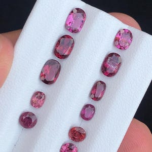 May include: An assortment of oval and round gemstones in varying shades of pink and red. The gemstones are displayed on a white surface, showcasing their polished facets and vibrant colors. The collection includes stones of different sizes.