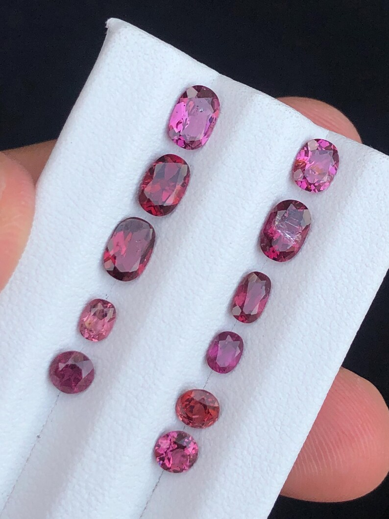 May include: An assortment of oval and round gemstones in varying shades of pink and red. The gemstones are displayed on a white surface, showcasing their polished facets and vibrant colors. The collection includes stones of different sizes.