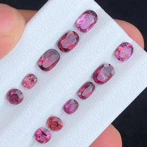 May include: A collection of oval and round gemstones in varying shades of pink and red, arranged on a white surface. The gemstones are displayed in a diagonal pattern, showcasing their polished surfaces and internal clarity. The gems are of different sizes.