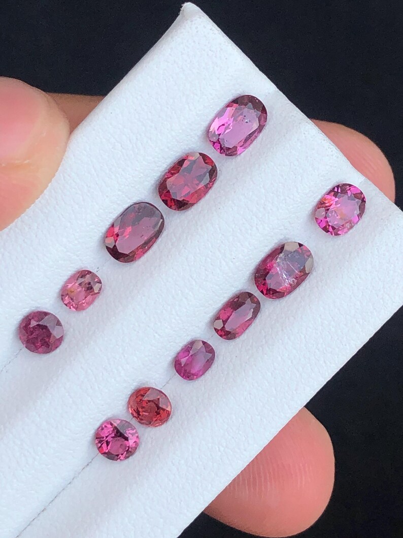 May include: A collection of oval and round gemstones in varying shades of pink and red, arranged on a white surface. The gemstones are displayed in a diagonal pattern, showcasing their polished surfaces and internal clarity. The gems are of different sizes.
