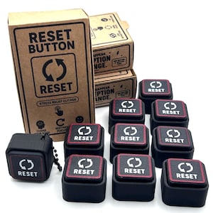 May include: A collection of black square stress relief clicker buttons with red trim and a white circular arrow icon above the word RESET. The buttons are displayed alongside brown cardboard packaging boxes featuring the text RESET BUTTON.