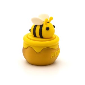 May include: A yellow and black bee figurine atop a yellow honey pot. The bee has black stripes and white wings. The honey pot has the word "HONEY" embossed on the side.