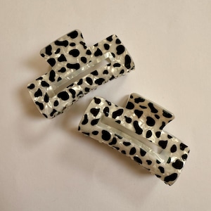 Puede incluir: Dos pinzas para el pelo rectangulares con un estampado de vaca. Las pinzas son blancas con manchas negras y tienen forma rectangular con la parte superior e inferior curvadas. Las pinzas están hechas de un material brillante.