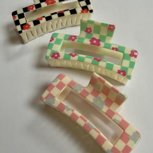 Puede incluir: Tres pinzas rectangulares para el cabello con patrones de tablero de ajedrez y detalles florales. Una pinza presenta un diseño a cuadros en blanco y negro con flores rojas, otra tiene un patrón a cuadros verde y blanco con flores rosas, y la última tiene un patrón a cuadros rosa y blanco con flores azules.