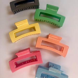 Puede incluir: Seis pinzas para el pelo de plástico en diferentes colores: gris, verde, amarillo, melocotón, rosa y azul. Las pinzas son todas del mismo tamaño y forma, con una abertura rectangular en el medio.