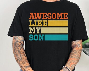 May include: Black t-shirt with the text "AWESOME LIKE MY SON" in orange, yellow, and teal. The shirt has a crew neck and short sleeves. The word "BLACK" is printed at the bottom right corner of the shirt.