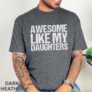 May include: Dark heather gray t-shirt with the text "AWESOME LIKE MY DAUGHTERS" in white. The shirt is a crew neck style and has short sleeves. The words "DARK HEATHER" are printed at the bottom of the image.